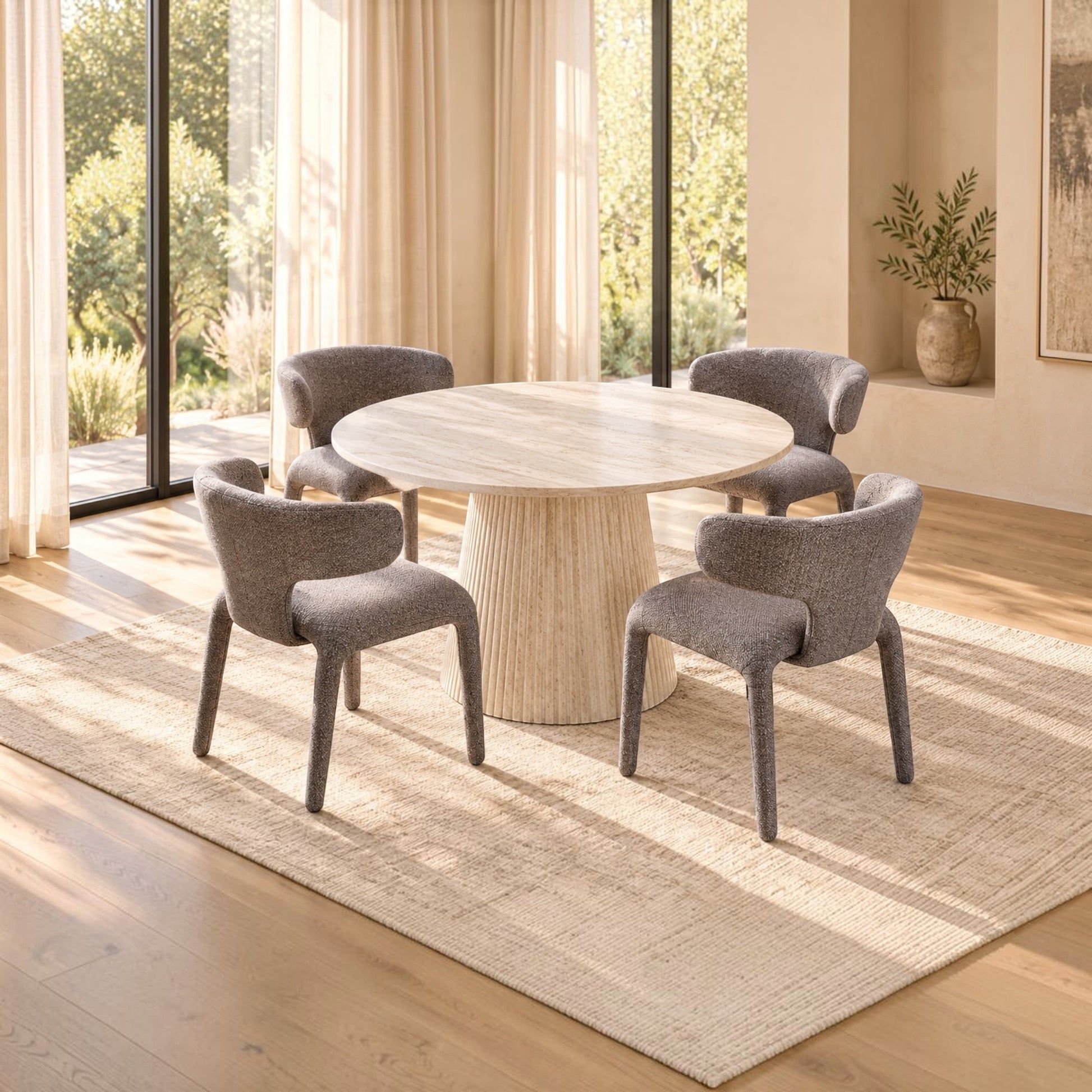 A round dining table with a textured cream pedestal base, surrounded by four upholstered dining chairs in a warm, modern dining room setting with large windows and a light beige rug.