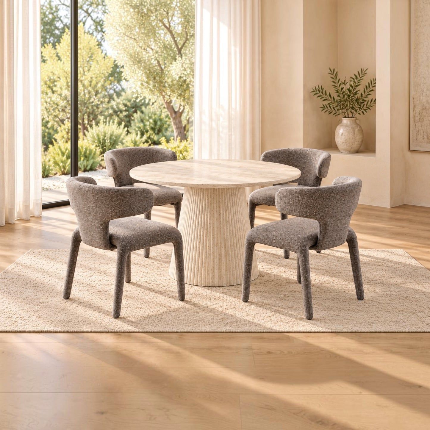 A modern dining set featuring a round light wood table and four upholstered gray chairs, arranged on a textured beige rug in a bright, contemporary dining room with large windows and natural light.