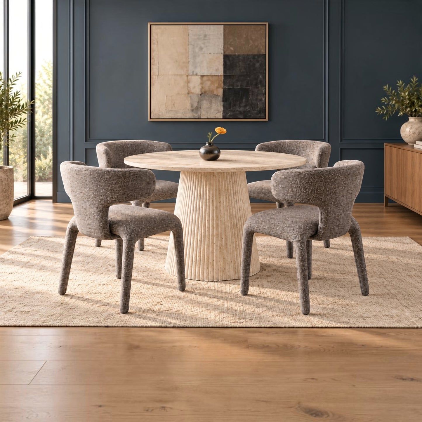 A round dining table with a ribbed pedestal base and four upholstered gray chairs, arranged on a light beige rug in a modern dining room with dark blue walls and wood flooring.