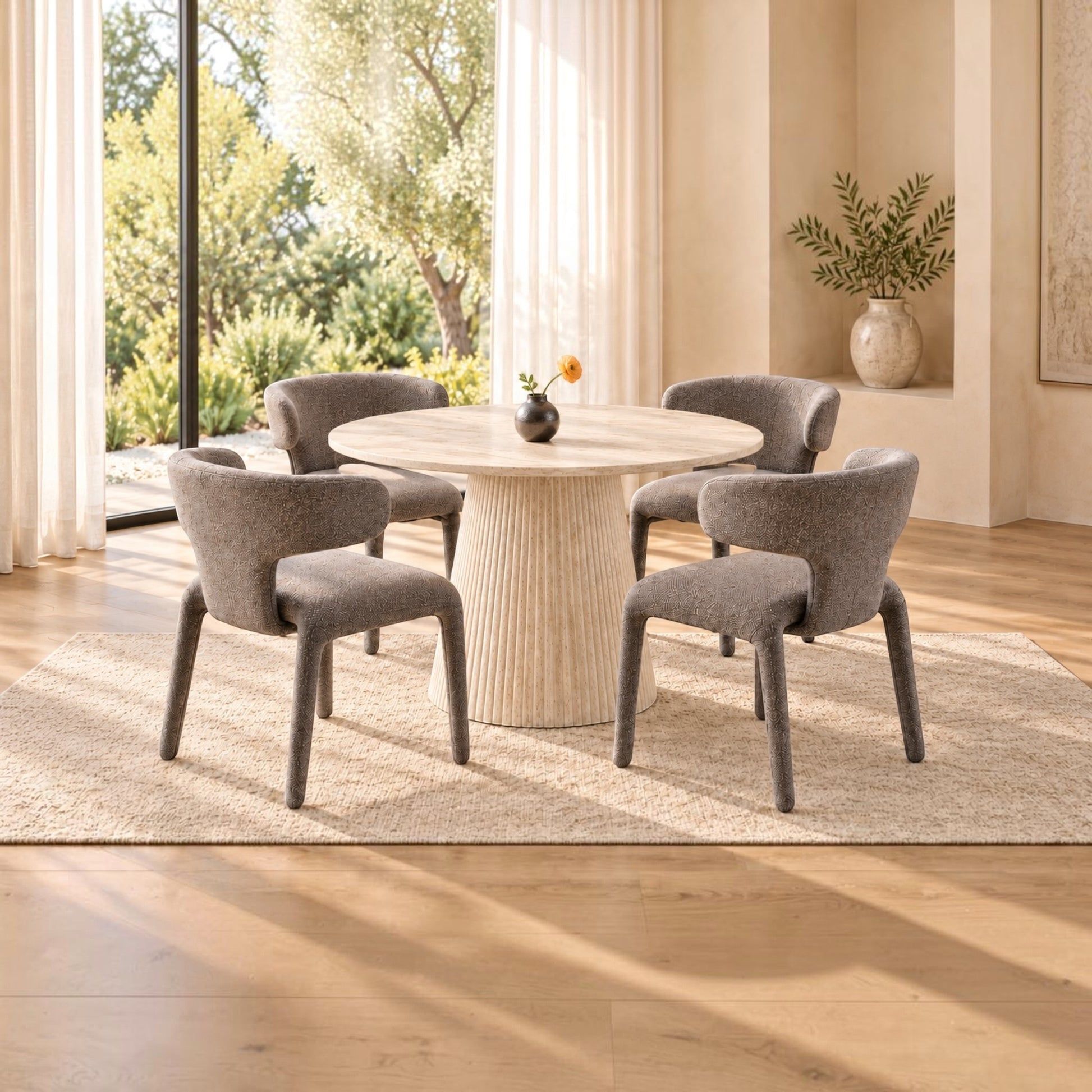 A modern dining set featuring a round ribbed pedestal table and four upholstered gray chairs, arranged on a light beige rug in a bright, sunlit room with large windows and a view of green trees.