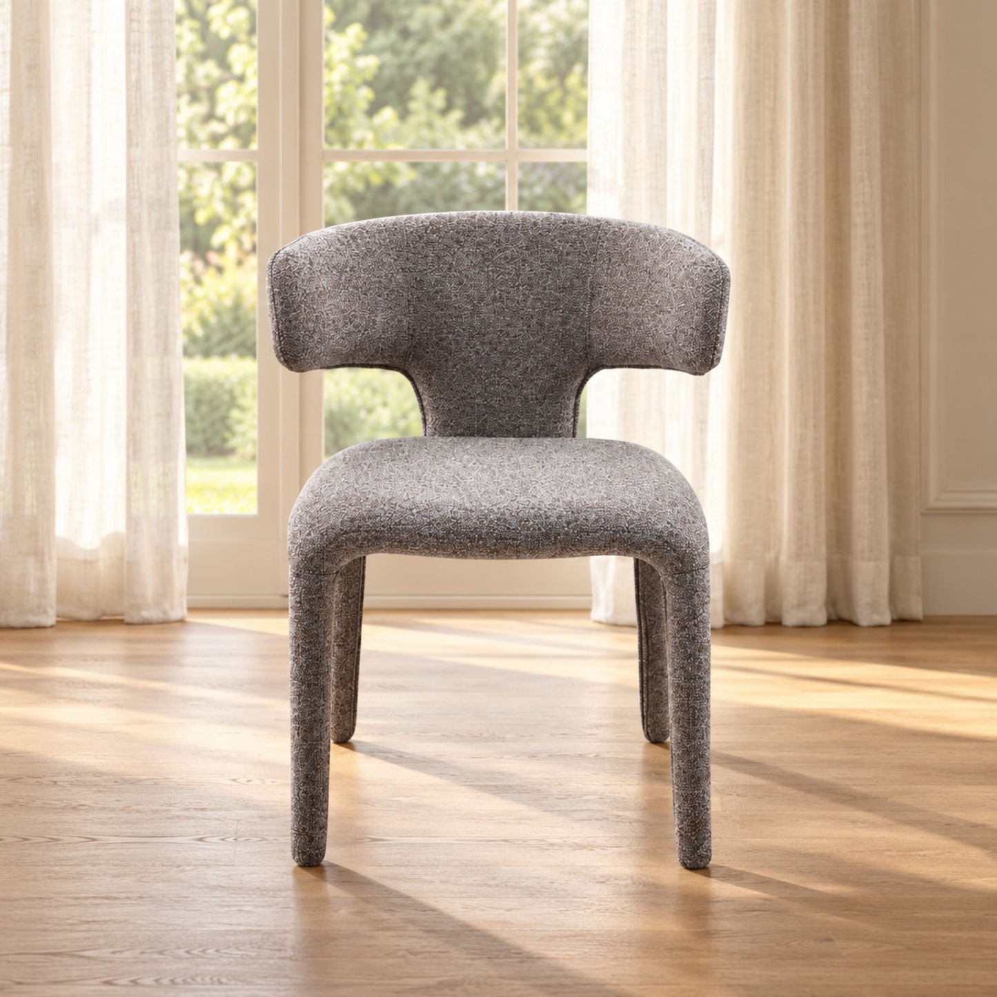A modern gray upholstered accent chair with a curved back and legs, positioned on a light wood floor in front of a window with sheer curtains.
