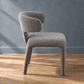 A modern upholstered dining chair with a curved backrest and textured boucle-style fabric in a warm gray-brown tone, shown on a wood floor against a blue-gray wall.