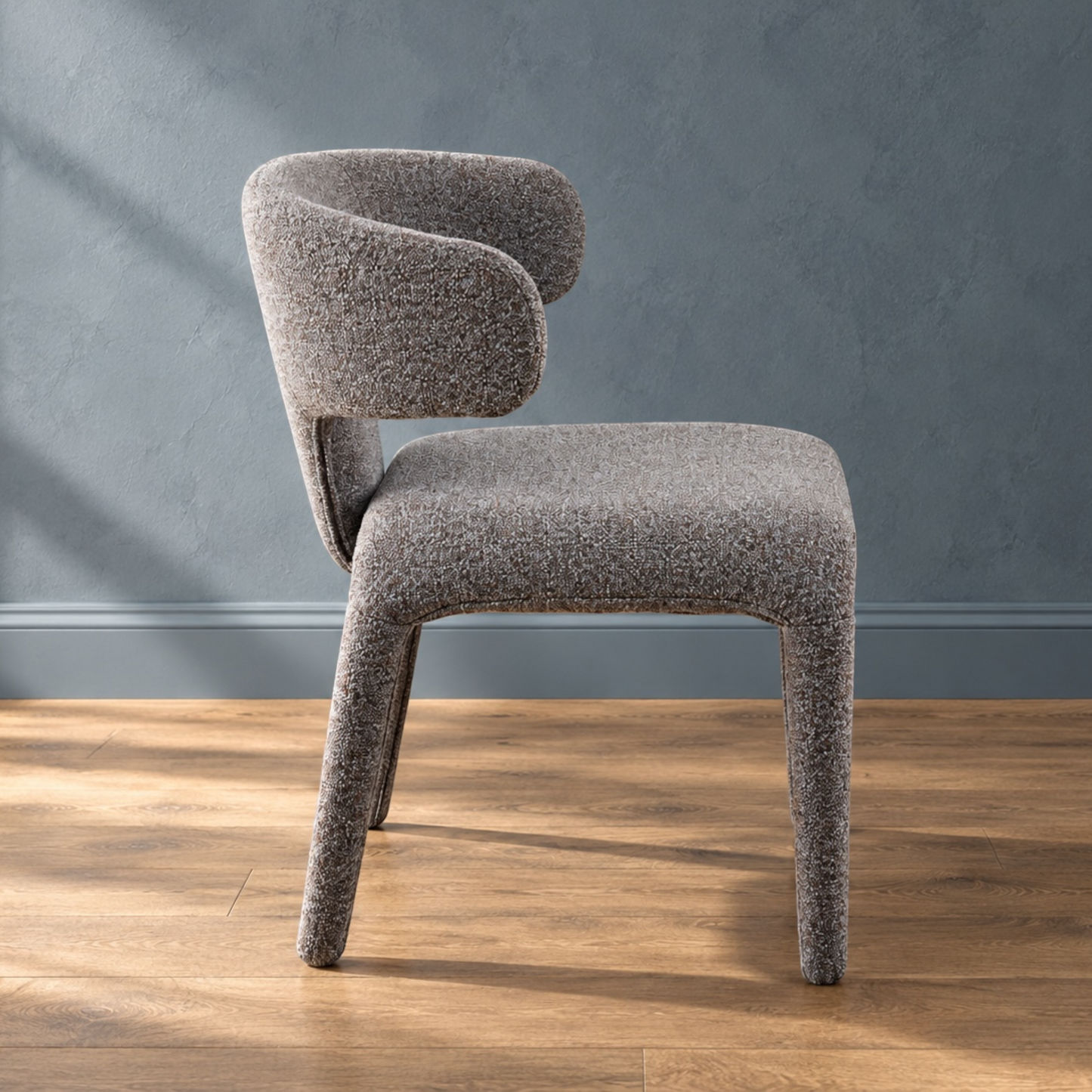 A modern upholstered dining chair with a curved backrest and textured boucle-style fabric in a warm gray-brown tone, shown on a wood floor against a blue-gray wall.