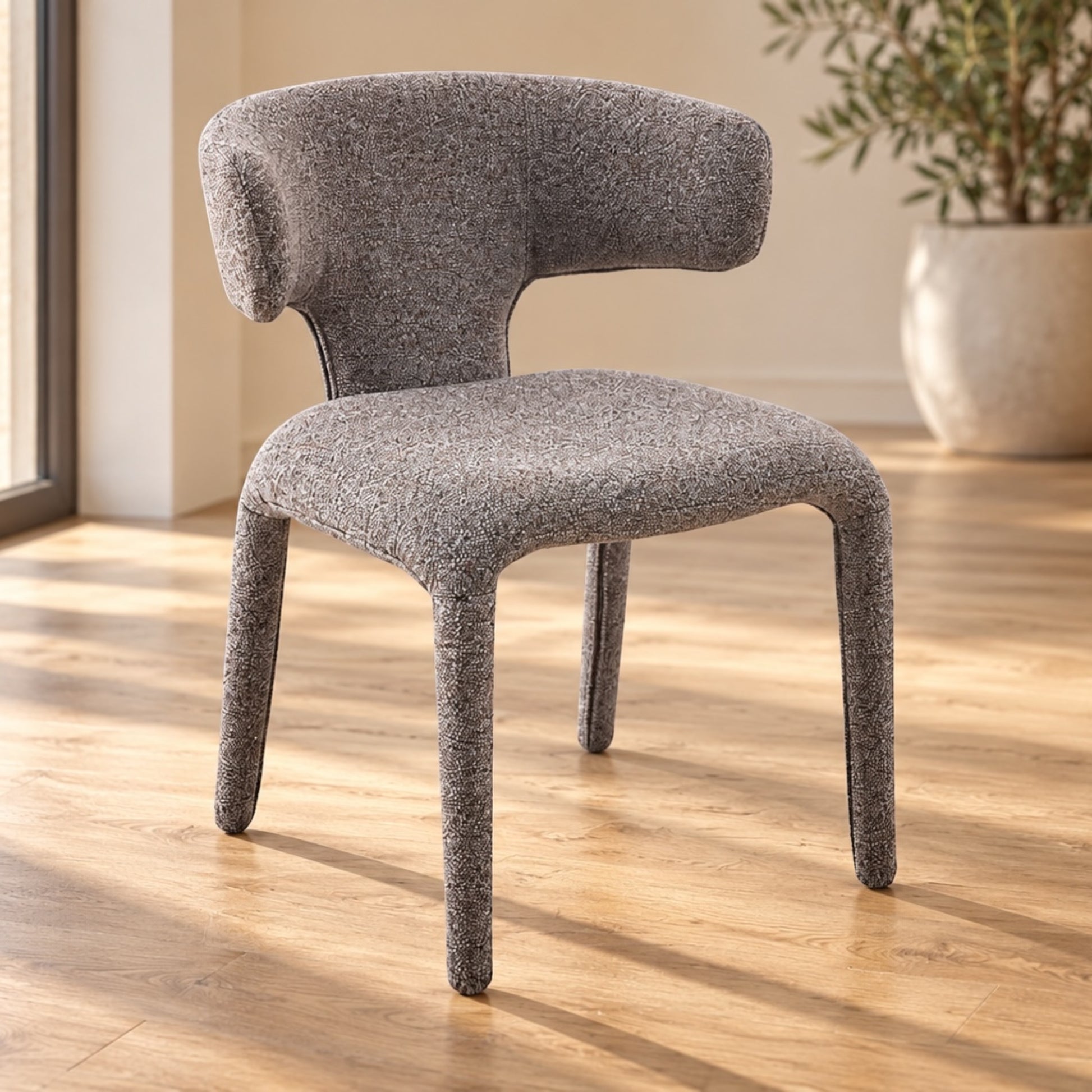 A modern upholstered dining chair with a curved backrest and legs, upholstered in a textured heather gray fabric, set on a light wood floor in a sunlit room.