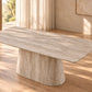 A modern rectangular dining table with a thick, rounded top in a light beige stone or woodgrain finish, supported by a wide, sculptural pedestal base, set in a warm, sunlit room with wood floors and neutral walls.