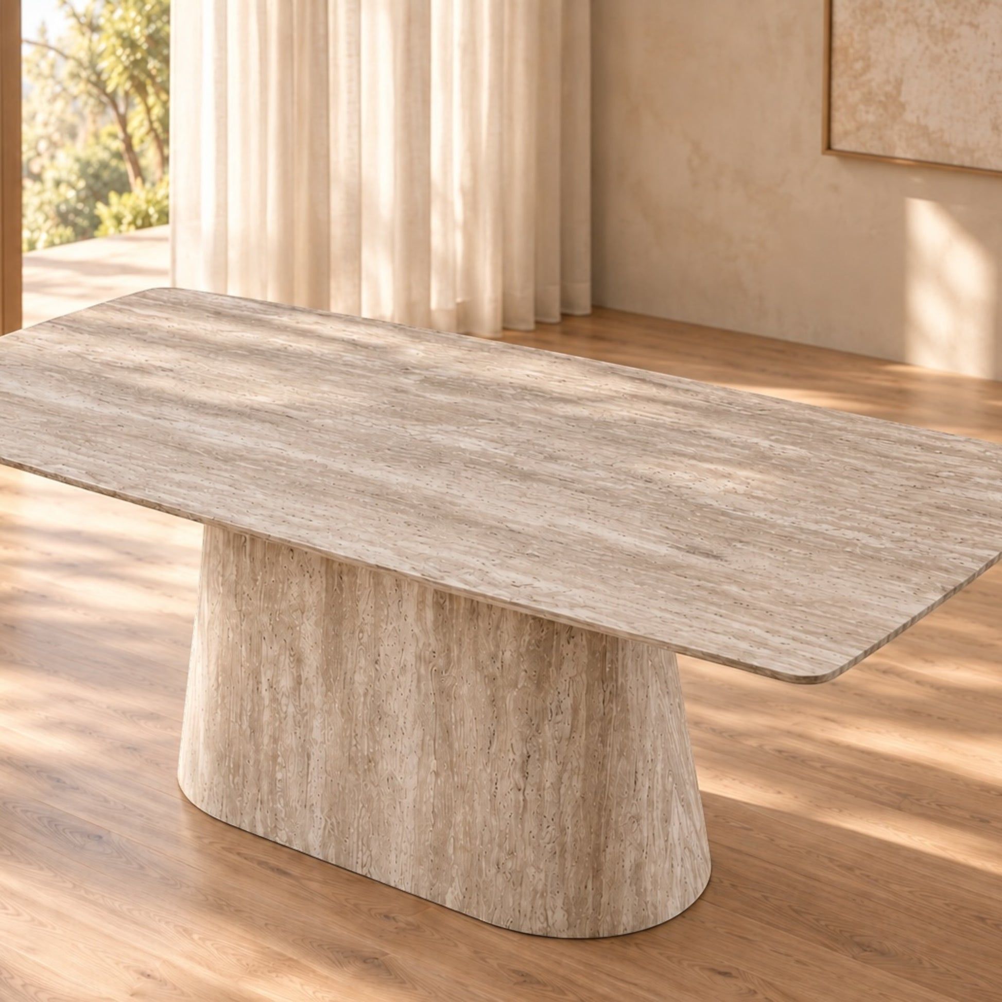 A modern rectangular dining table with a thick, rounded top in a light beige stone or woodgrain finish, supported by a wide, sculptural pedestal base, set in a warm, sunlit room with wood floors and neutral walls.