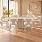 A modern dining set featuring a rectangular stone table and six matching upholstered chairs in a warm, sunlit room with wood floors and beige walls.