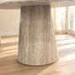 A close-up of a round dining table with a thick, fluted pedestal base in a light weathered gray stone or concrete finish, set on a light wood floor in a bright room.