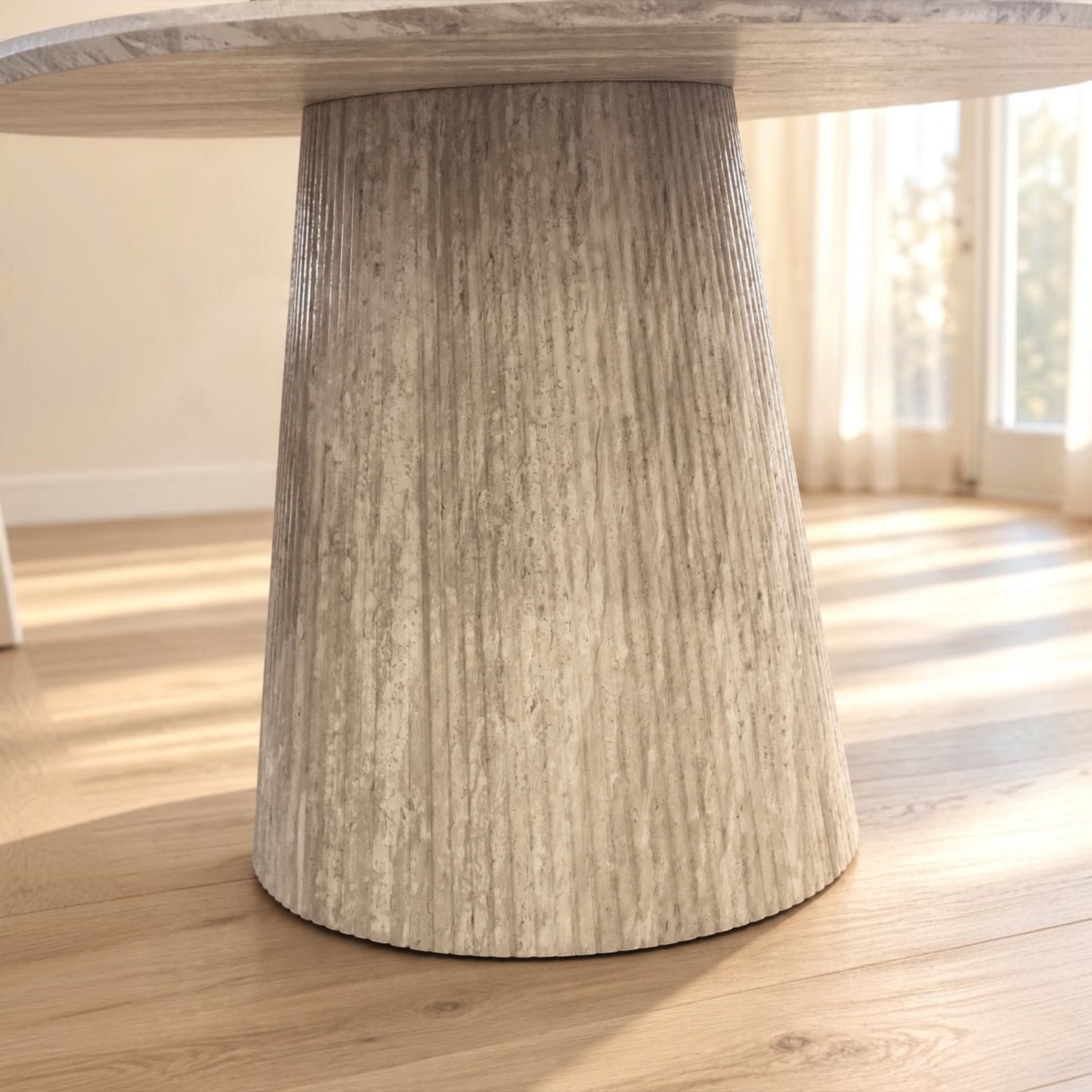 A close-up of a round dining table with a thick, fluted pedestal base in a light weathered gray stone or concrete finish, set on a light wood floor in a bright room.