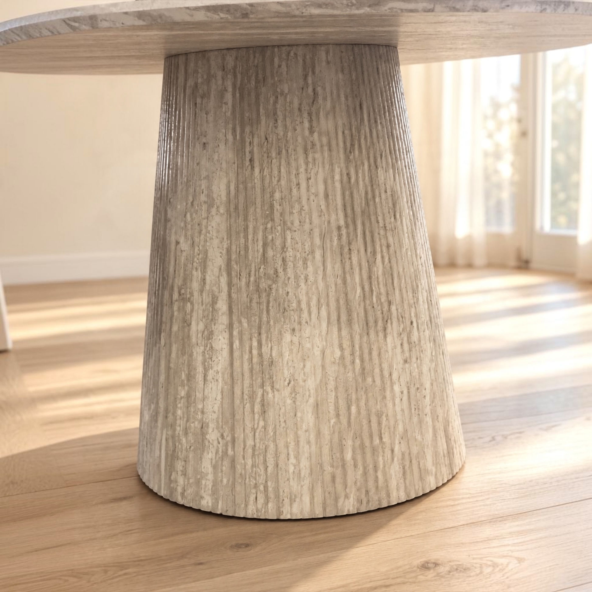 A close-up of a round dining table with a thick, fluted pedestal base in a light weathered gray stone or concrete finish, set on a light wood floor in a bright room.