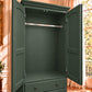 A dark green wooden armoire or wardrobe with two doors open, showing an interior rod and shelf, set in a warm-toned room with a window and curtains.