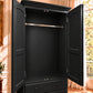 A tall black armoire or wardrobe cabinet with two open doors, a metal hanging rod, and a lower drawer, shown in a real interior room with warm sunlight and window light.