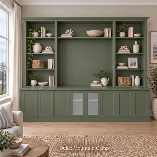 A large built-in shelving unit in a muted sage green color, styled with books, ceramic vases, baskets, and green plants in a bright living room. Text at the bottom reads 'Dulux Mountain Camp'.