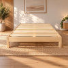 A light NZ pine wood slatted bed frame shown in a styled bedroom setting with a woven jute rug, wooden floor, potted plants, and wall art.