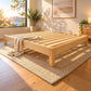 A light NZ pine wood platform bed frame with slatted support bars, shown in a bright, sunlit bedroom setting on a woven rug with a throw blanket draped over the corner.