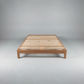 A minimalist wooden bed frame with a slatted platform base, shown from a front-three-quarters angle against a neutral gray studio background.