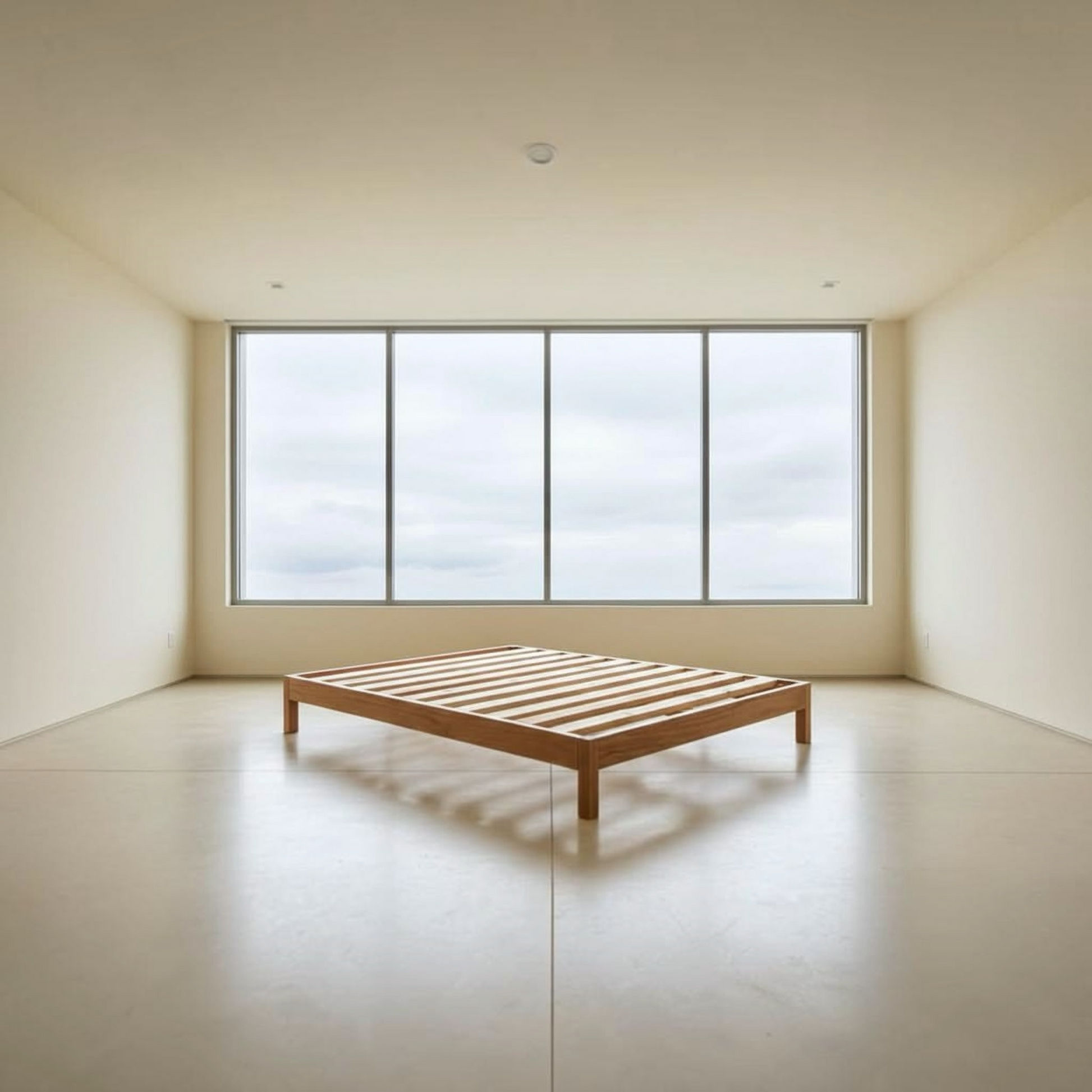 A minimalist wooden slat bed frame in a warm medium brown tone, positioned in a spacious, modern room with cream walls, a polished concrete floor, and a large multi-pane window looking out to a cloudy sky.