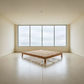 A minimalist wooden slat bed frame in a warm medium brown tone, positioned in a spacious, modern room with cream walls, a polished concrete floor, and a large multi-pane window looking out to a cloudy sky.