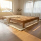 A minimalist wooden platform bed frame with slatted support beams, styled in a bright, sunlit room with a cream shag rug and a draped throw blanket.