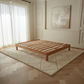A light natural wood platform bed frame with slatted support bars, shown in a bright, modern bedroom setting on a cream-colored textured rug over a light wood herringbone floor.