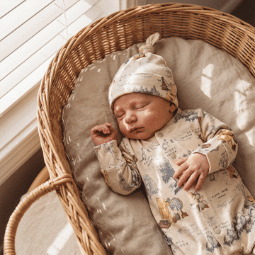 A sleeping newborn baby wearing a beige Winnie the Pooh hat and matching long-sleeve romper, resting in a woven wicker bassinet with a cushioned fabric liner.