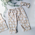 Flat lay image of a pair of kids baggy, beige trousers with white flower print. Pictured with a coordinating knotted headband. 
