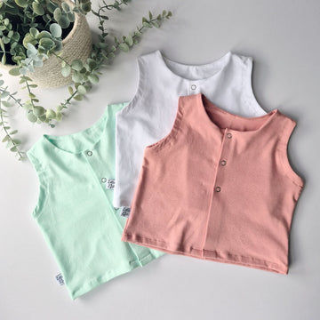 Three sleeveless shirts in green, white, and pink on a light gray surface with a plant in the background.