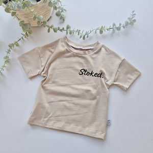 Beige t-shirt with 'Stoked' text on a light background with greenery