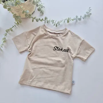 Beige t-shirt with 'Stoked' text on a light background with greenery