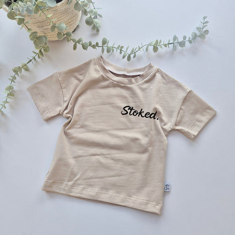 Beige t-shirt with 'Stoked' text on a light background with greenery