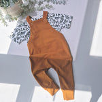 Brown overalls on a light gray surface with a plant and patterned fabric in the background.