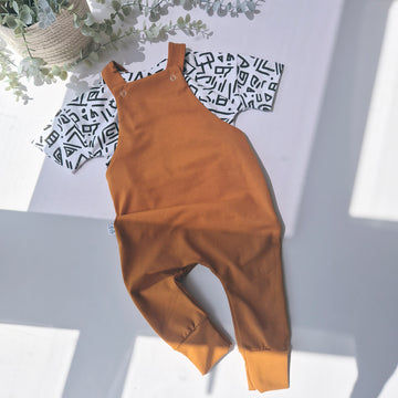 Brown overalls on a light gray surface with a plant and patterned fabric in the background.