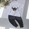 White t-shirt with black graphic and dark pants on a light surface with plants in the background