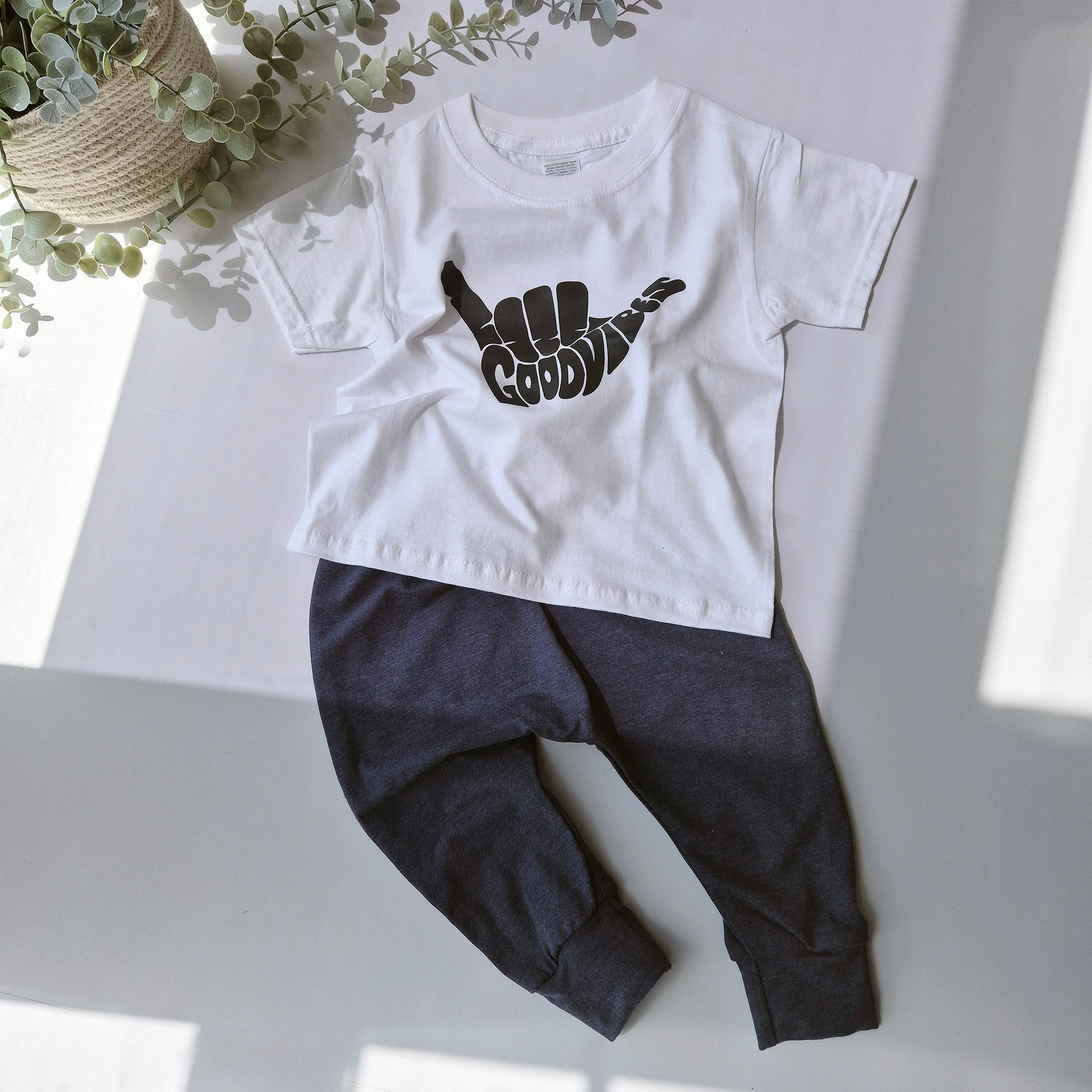 White t-shirt with black graphic and dark pants on a light surface with plants in the background