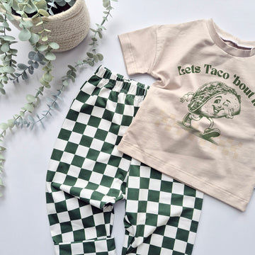 Children's t-shirt with graphic design and green checkered pants on a light surface with plants.