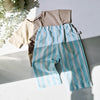 Children's clothing including a beige shirt and blue striped pants on a light surface with a plant in the background.