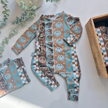 Baby onesie with smiley face and peace sign pattern on a white surface with greenery.