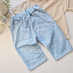 Light blue denim look trousers on a beige surface with a beige shirt and plant in the background.