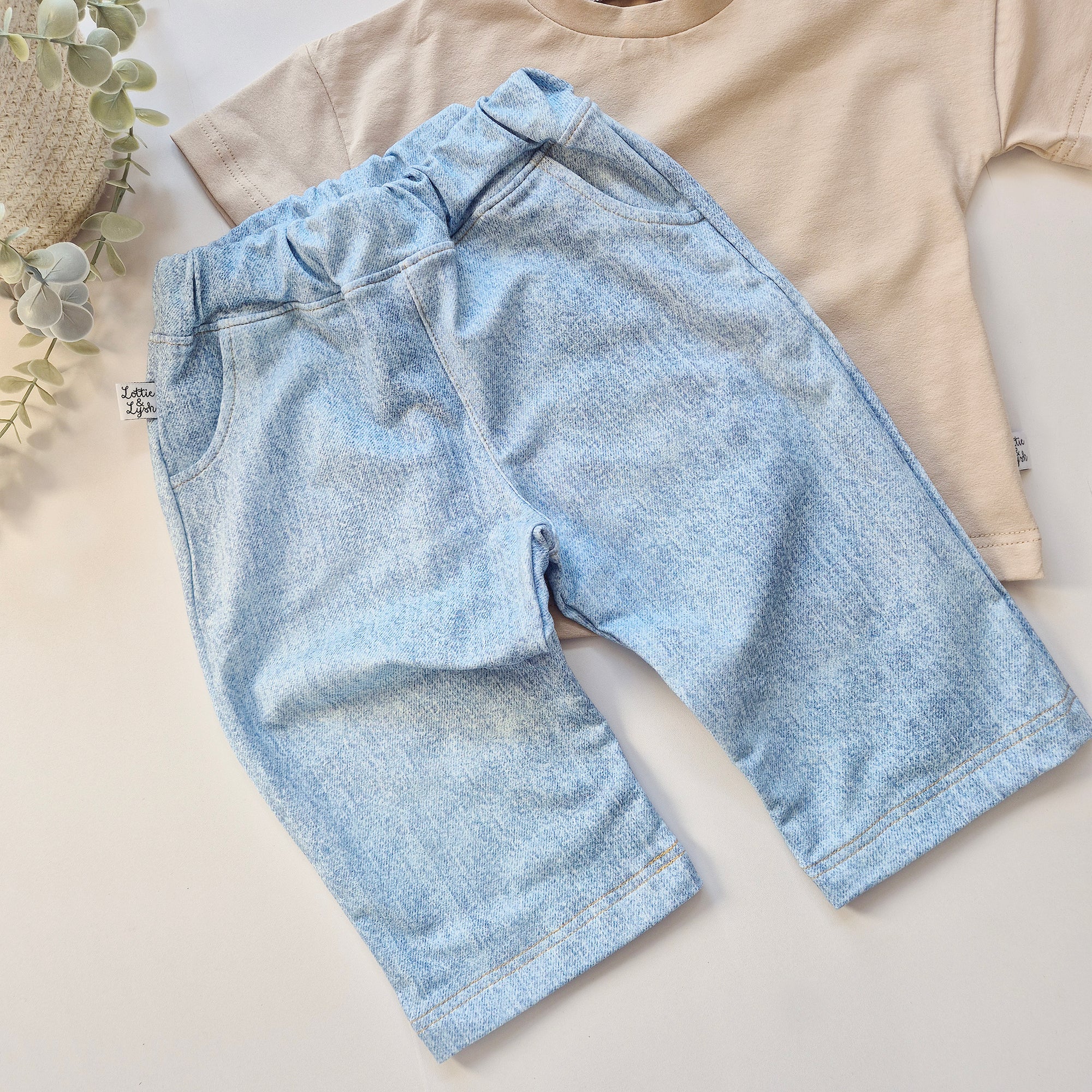Light blue denim look trousers on a beige surface with a beige shirt and plant in the background.