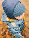 Child wearing a blue coat with leaf pattern and navy hat, standing in autumn leaves.