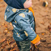 Child wearing a blue jacket with yellow accents and a hood, standing on a dirt path with autumn leaves.