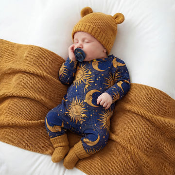 Baby sleeping in a blue onesie with star and moon patterns, wearing a mustard yellow knit hat and matching booties, on a brown blanket.