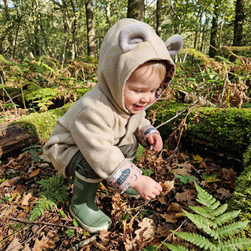 Child in a forest wearing a bear-themed hoodie and green boots.