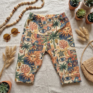 Boho-style baby pants with a colorful bohemian print featuring suns, eyes, palm trees, and coral, laid flat on a textured white fabric with decorative props including sunglasses, a wooden bead necklace, potted plants, and a woven bag.