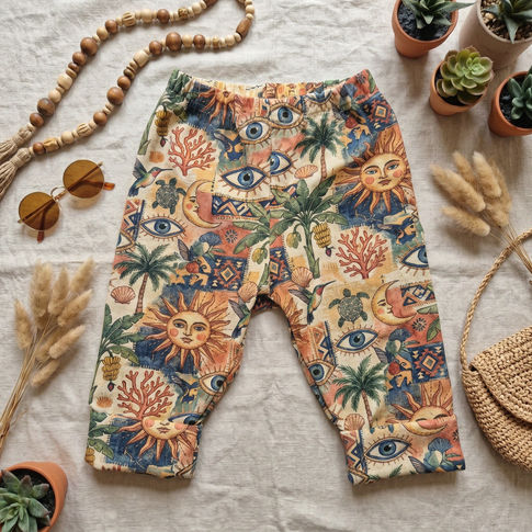 Boho-style baby pants with a colorful bohemian print featuring suns, eyes, palm trees, and coral, laid flat on a textured white fabric with decorative props including sunglasses, a wooden bead necklace, potted plants, and a woven bag.