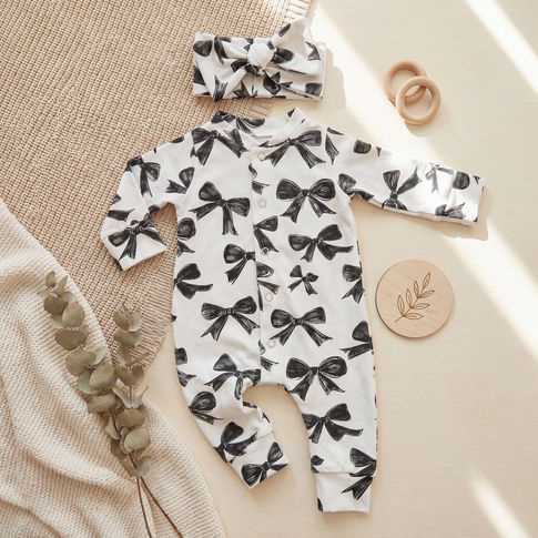 A baby outfit set featuring a white long-sleeve footless romper with a black bow print, a matching headband, and wooden baby accessories, arranged in a styled flat-lay on a neutral background.