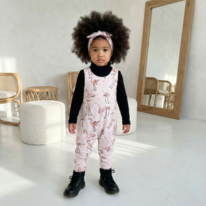 Young child wearing a pink flamingo print dungarees with black boots in a minimalistic room.