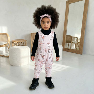 Young child wearing a pink flamingo print dungarees with black boots in a minimalistic room.