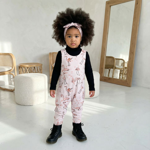 Young child wearing a pink flamingo print dungarees with black boots in a minimalistic room.