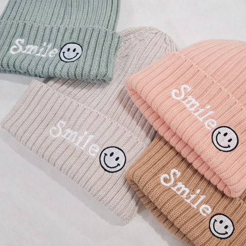 Four knit hats in different colors with 'Smile' text and smiley face icons on a light background.